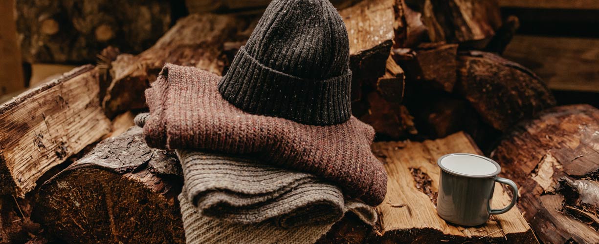 A gray knit hat and two folded sweaters are stacked on firewood next to a gray enamel mug; the setting appears rustic and cozy, suggesting warmth and comfort.