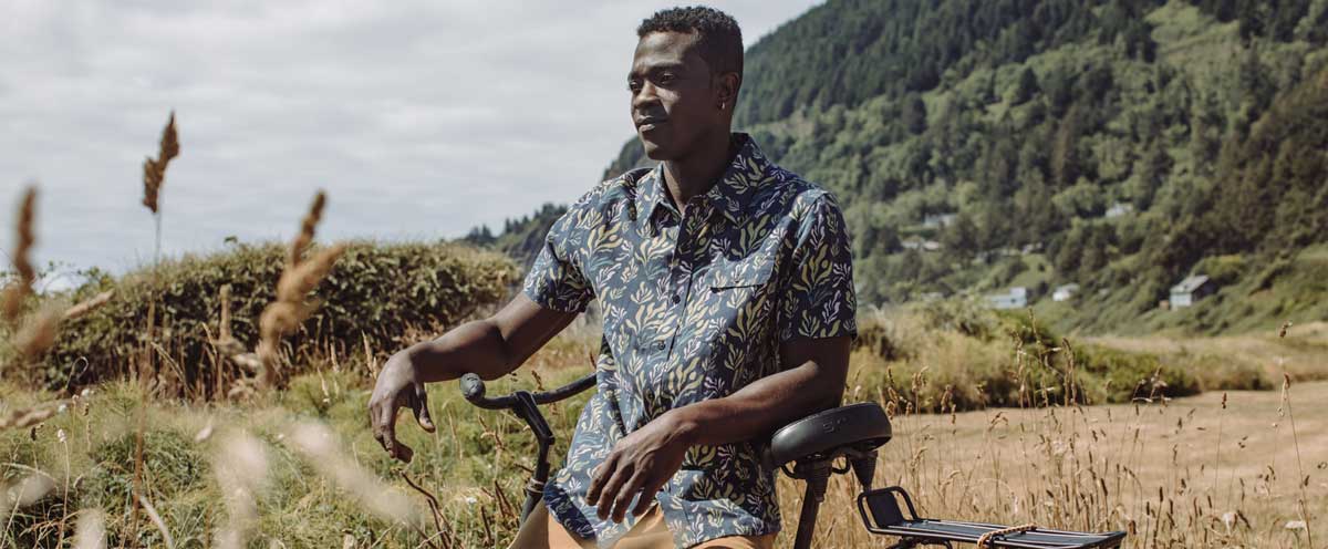 A man wearing a floral shirt sits on a bicycle in a grassy field with green, forested hills and cloudy sky in the background.