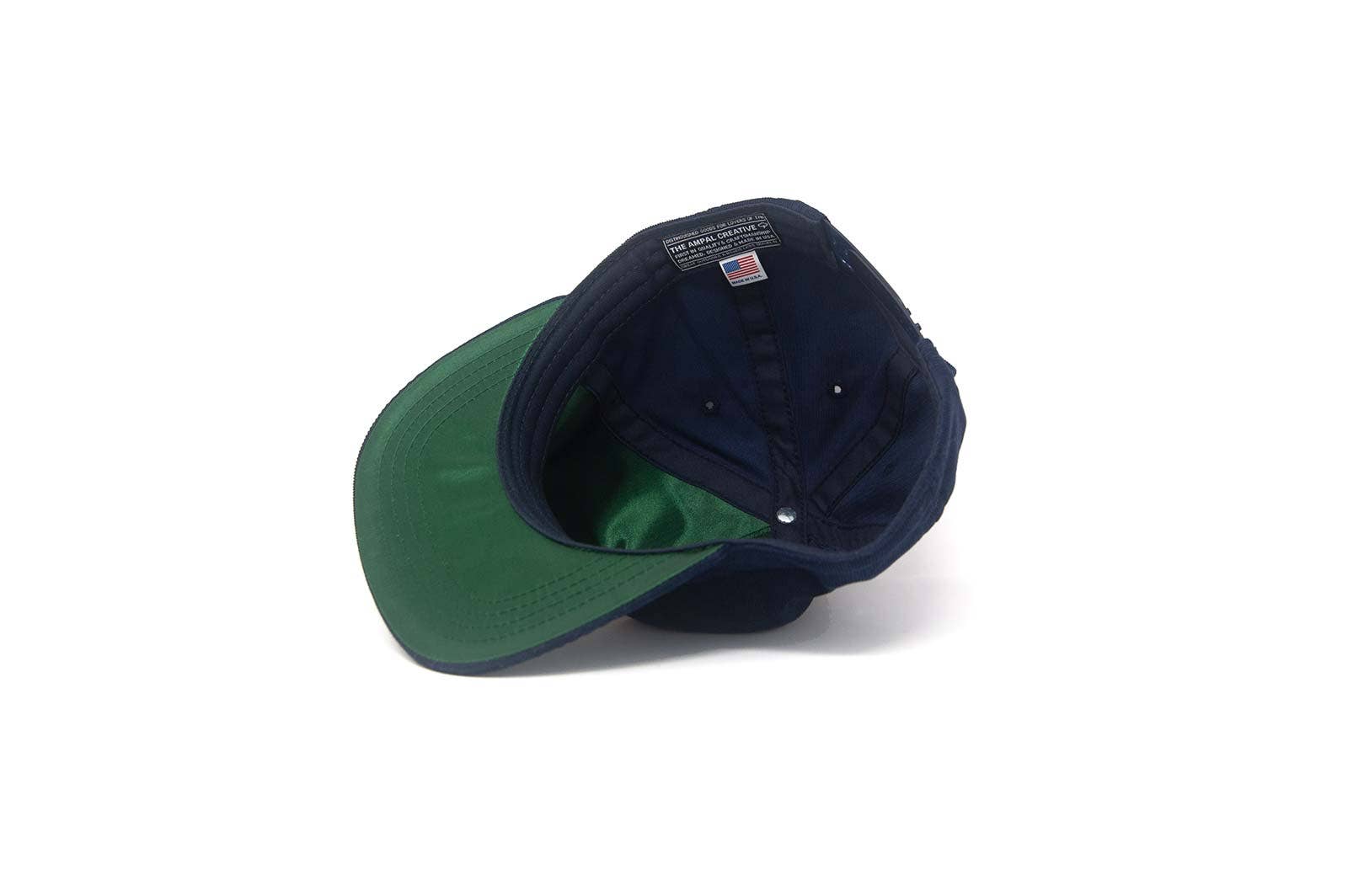 The Ampal Creative - Tiger III Snapback in navy is shown upside down on a white background, highlighting its green underbill, black sweatband, and an interior label with an American flag.
