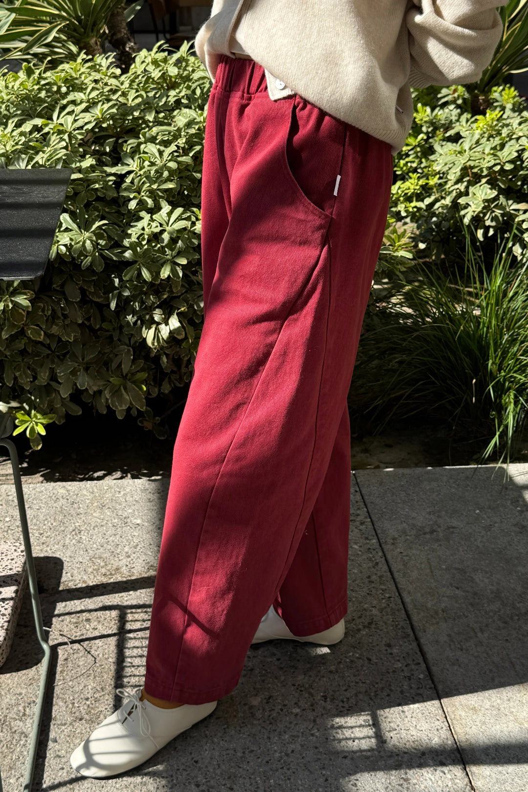 A person wears dark red, cropped-length Le Bon Shoppe - Arc Pant / Oxblood with an elastic waist and side pockets, paired with a cream-colored sweater and white shoes.