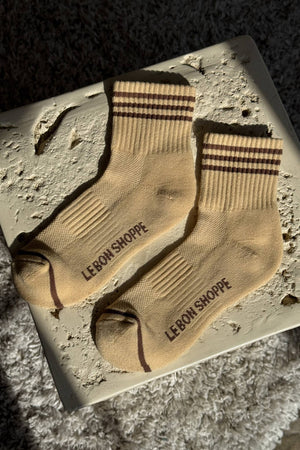 A pair of Le Bon Shoppe - Girlfriend Socks / Daisy in beige with brown stripes and "LE BON SHOPPE" on the side, laid flat on a textured cream surface in sunlight.