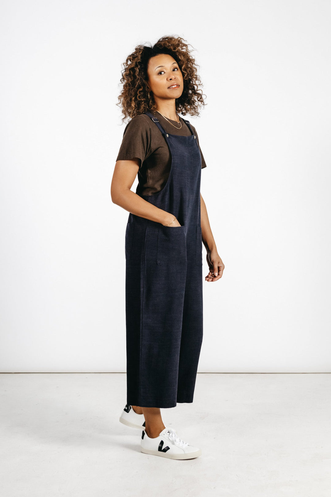A woman with curly hair stands confidently against a white background, wearing the Bridge & Burn Edie Overall in Navy—a wide-leg, linen-cotton blend piece with adjustable straps—paired with a brown T-shirt and her hands in her pockets.