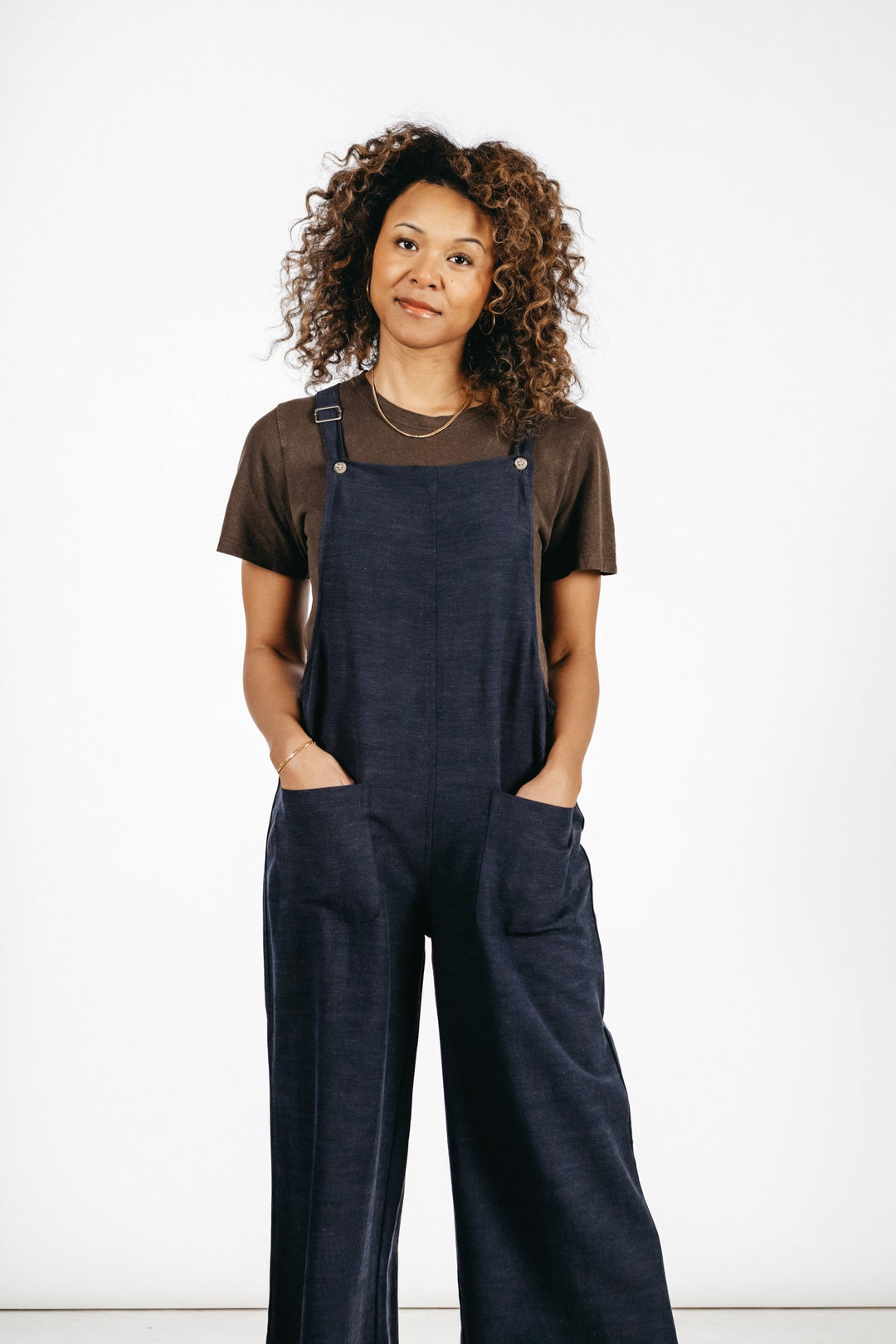 A person with curly hair wears a brown t-shirt and the Bridge & Burn Edie Overall in Navy, featuring adjustable straps and wide legs, standing hands-in-pockets against a plain white background.