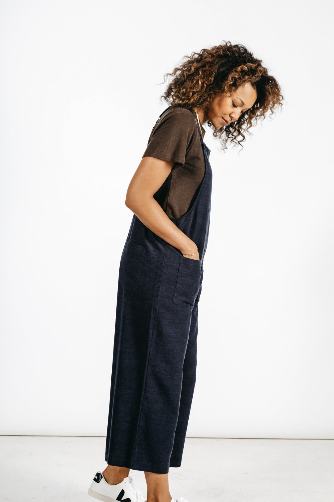 A woman with curly hair stands in profile against a white background, wearing Bridge & Burn’s Edie Overall in Navy over a dark brown T-shirt and white sneakers. Hands in her pockets, she looks down and smiles softly.