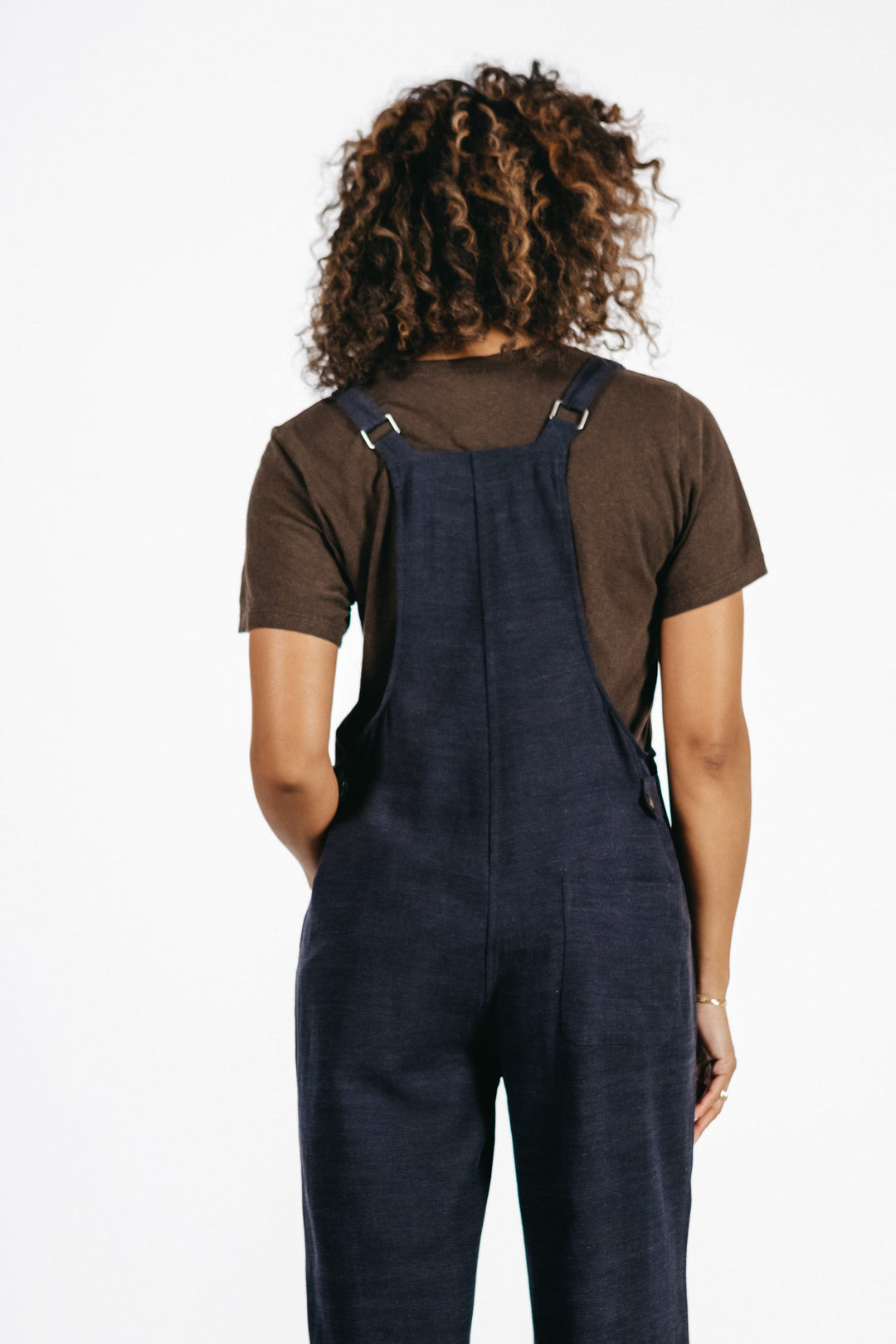 A person with curly hair, facing away, wears the Bridge & Burn Edie Overall in Navy with a brown t-shirt against a plain white background.
