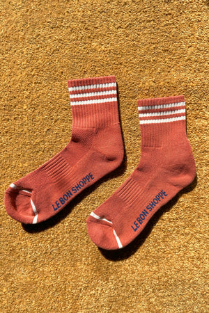 A pair of Le Bon Shoppe - Girlfriend Socks in Terracotta with white stripes at the top and blue branding on the soles, displayed on a brown textured carpet in sunlight.