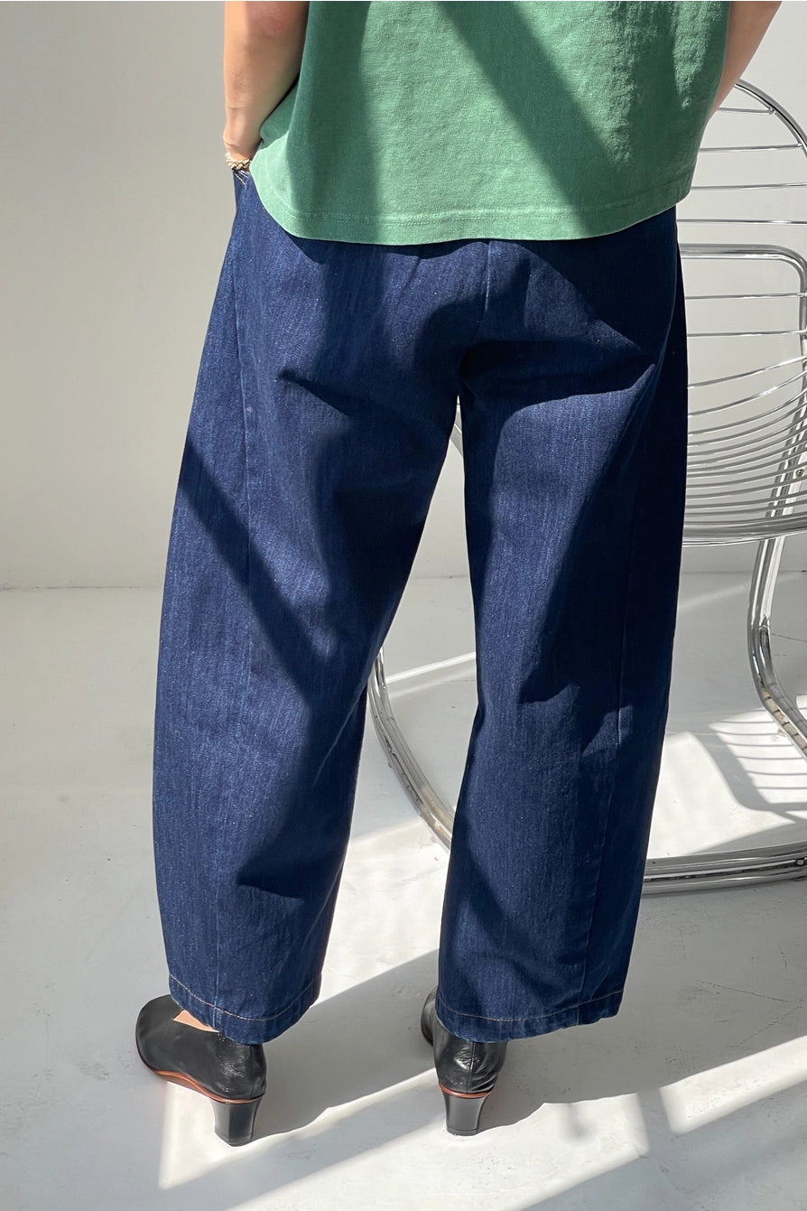 A person wearing a green shirt, Le Bon Shoppe’s Arc Pant in Blue Denim, and black shoes stands by a metal chair on a light floor with sunlight casting shadows.