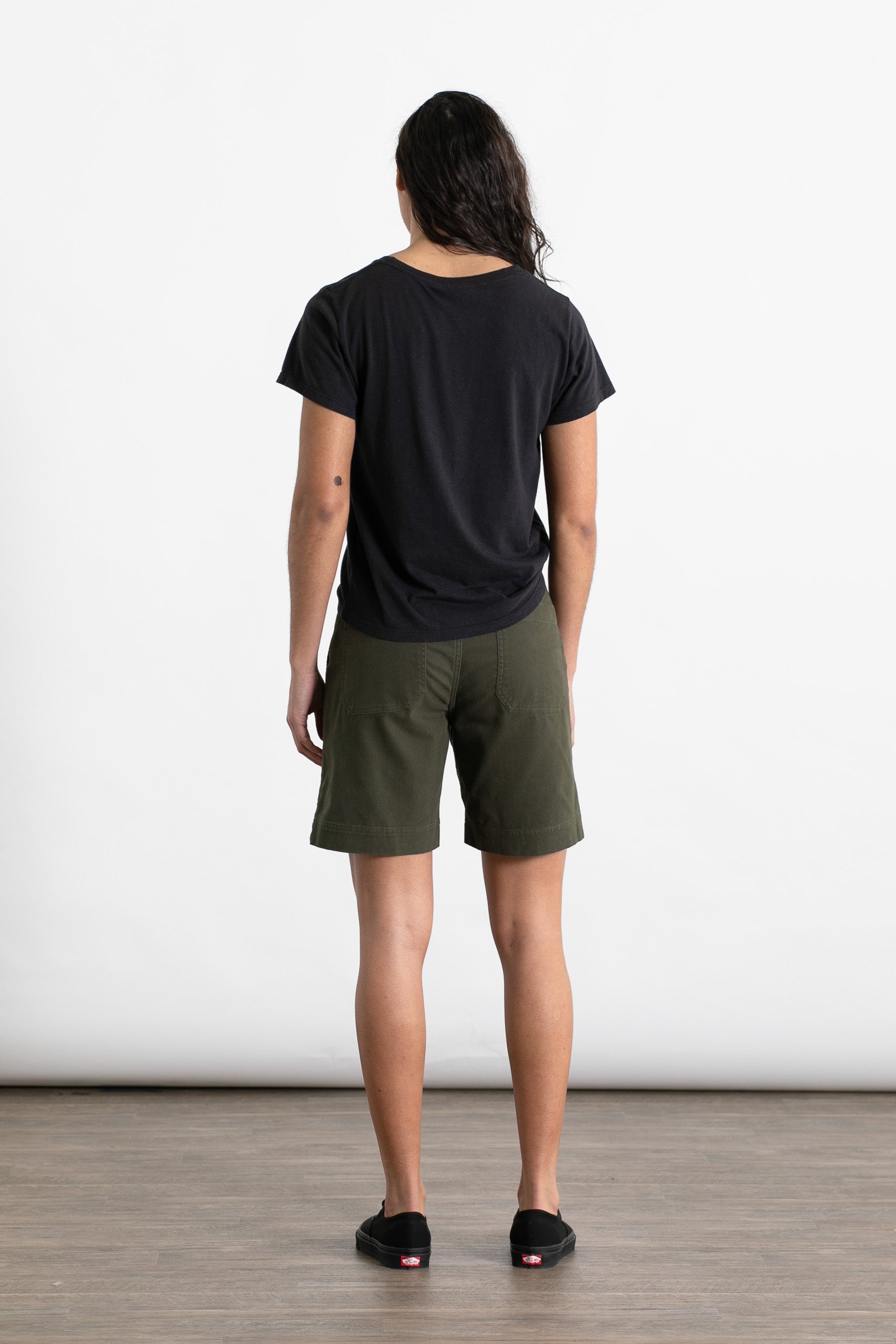 A person with long dark hair, wearing the Jungmaven Ojai Tee in Black, stands facing away from the camera on a wooden floor with a white background.