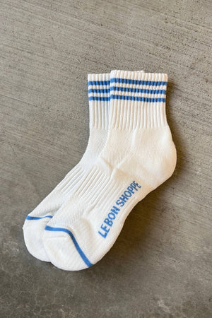 A pair of Le Bon Shoppe - Girlfriend Socks in Ivory with two blue cuff stripes, blue toe trim, and "LE BON SHOPPE" printed in blue on the sole, shown on a concrete surface.