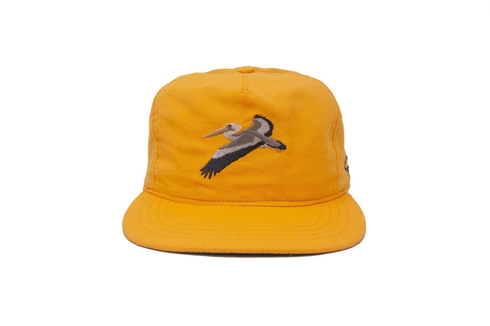 The Ampal Creative - Glider Strapback / Gold: A water-repellent, lightweight nylon hat in bright yellow with an embroidered flying pelican on the front. Shown against a plain white background.