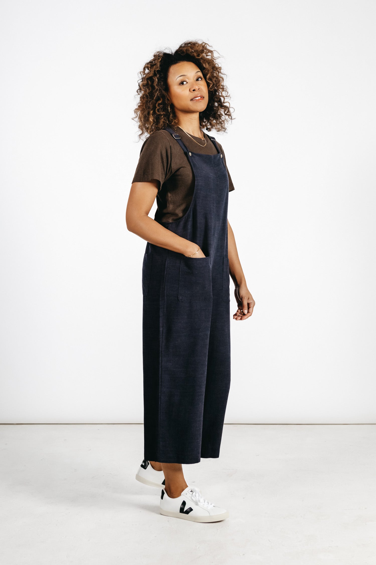 A woman with curly hair wears a brown t-shirt, white sneakers, and the Bridge & Burn Edie Overall in Navy—a wide-leg linen-cotton blend overall with adjustable straps—standing in a minimalist studio with a white background.