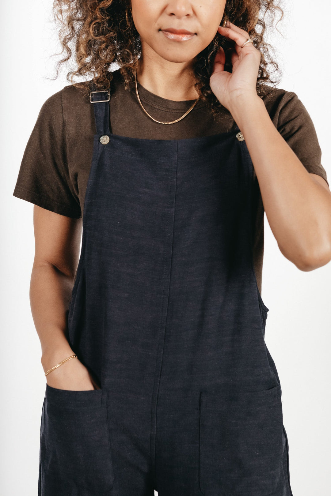 A woman wearing the Bridge & Burn Edie Overall in Navy, featuring adjustable straps and a wide leg silhouette, stands with one hand in her pocket and the other touching her hair against a plain white background.