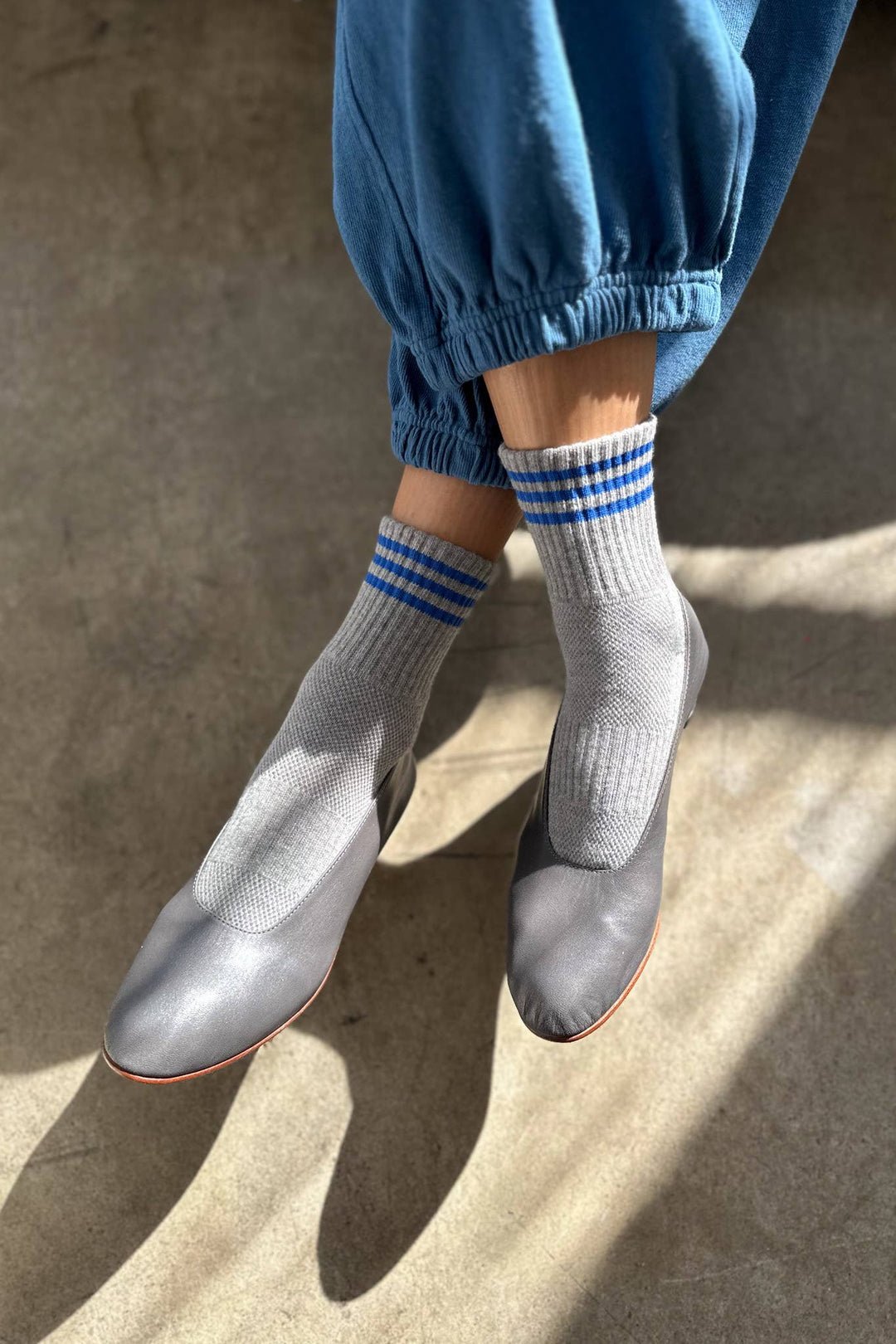 A person wearing Le Bon Shoppe Girlfriend Socks in grey stands on a sunlit concrete floor, paired with gray slip-on shoes and blue cuffed pants.