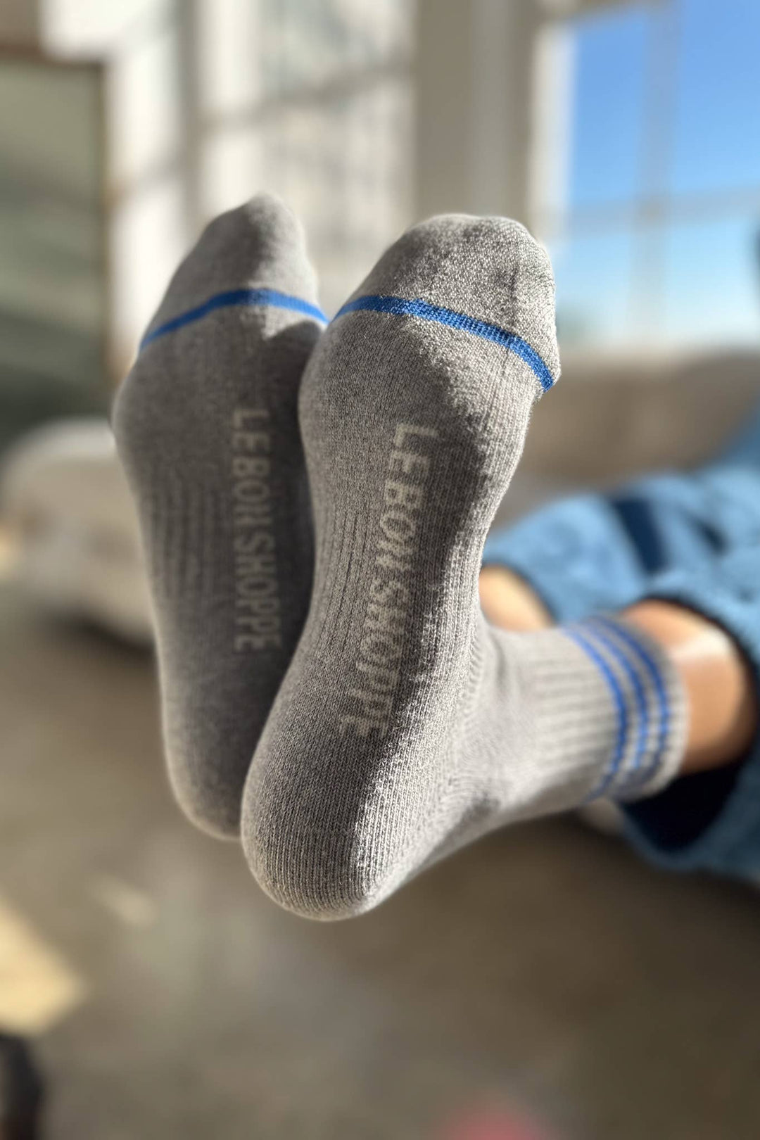 A person wearing Le Bon Shoppe - Girlfriend Socks in grey with blue stripes rests with feet together, showing "LEBONSHOPPE" in white on the soles as sunlight streams through a window.
