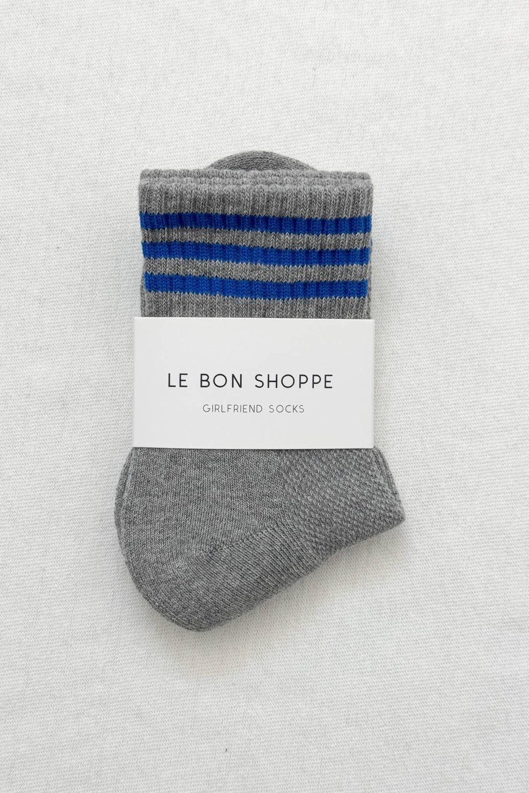 A pair of Le Bon Shoppe - Girlfriend Socks in grey, featuring two blue stripes near the top, folded with a white label that reads "LE BON SHOPPE Girlfriend Socks," displayed on a light textured surface.