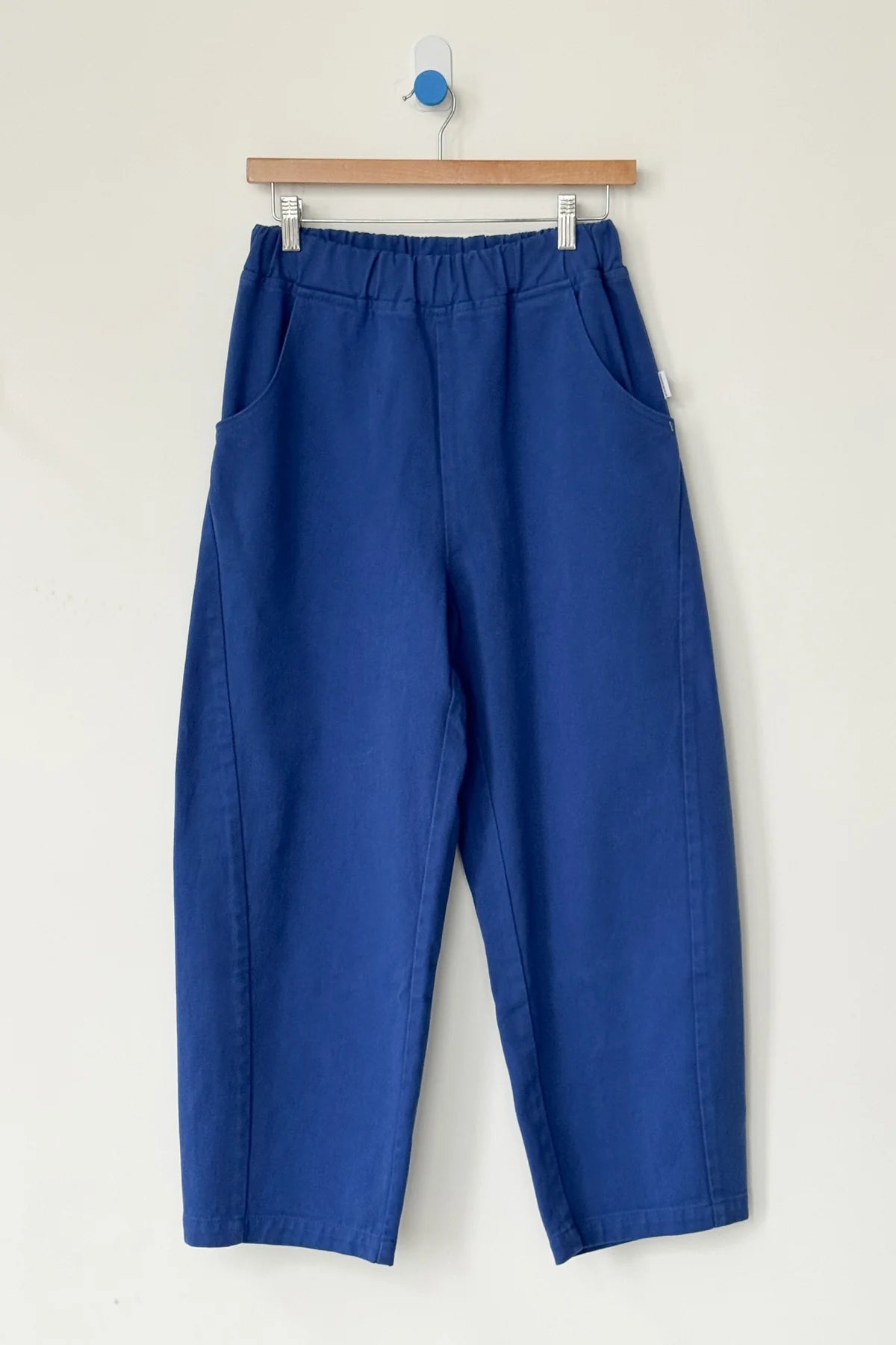 Le Bon Shoppe Arc Pant in Azure: a pair of loose-fitting, high-waisted pants with an elastic waistband and side pockets, crafted from heavyweight denim. Shown hanging on a wooden hanger against a light wall.