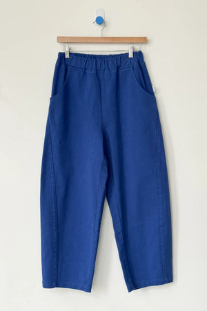 Le Bon Shoppe Arc Pant in Azure: a pair of loose-fitting, high-waisted pants with an elastic waistband and side pockets, crafted from heavyweight denim. Shown hanging on a wooden hanger against a light wall.