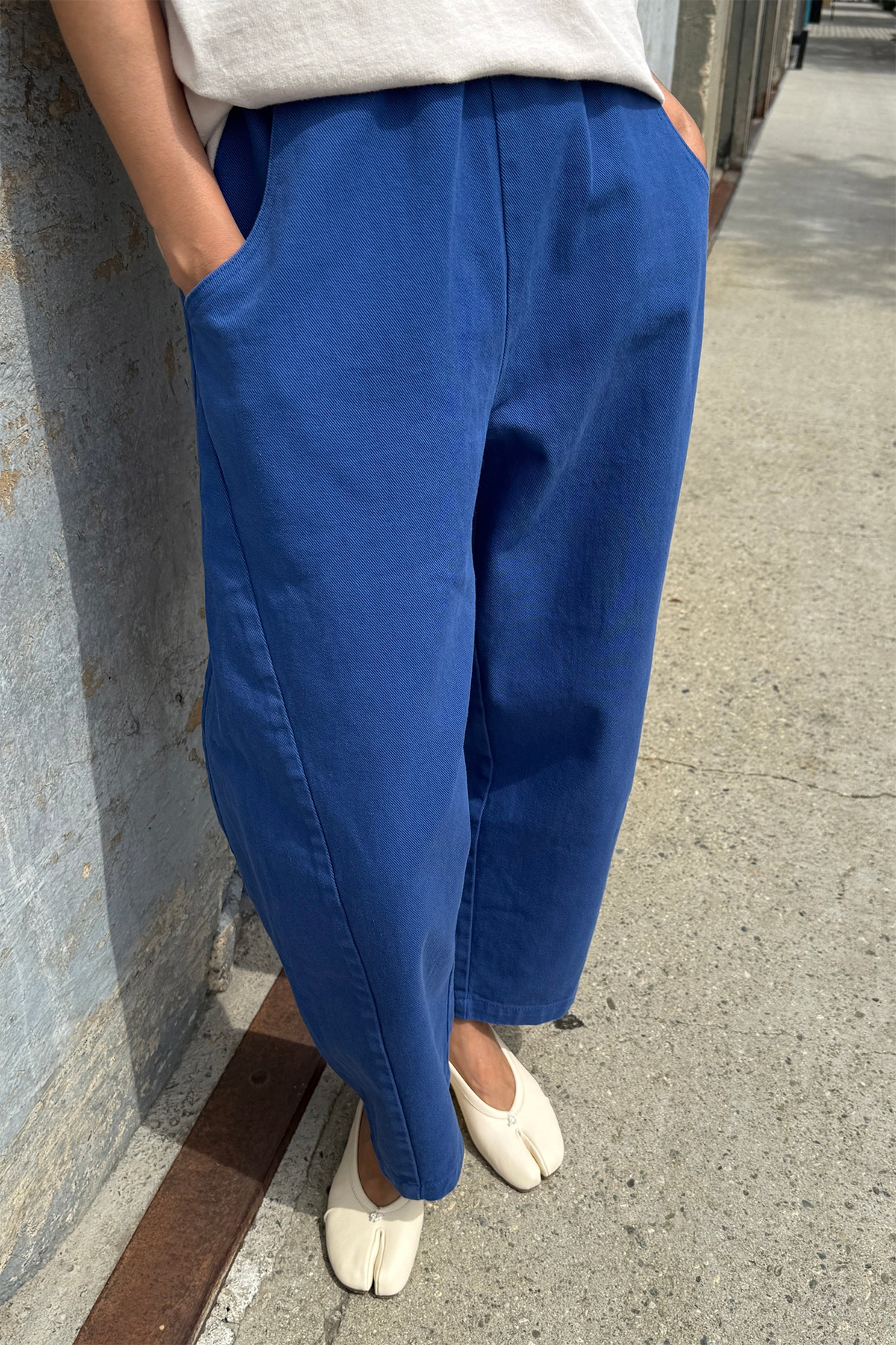 A person stands outdoors on a sunny day in Le Bon Shoppe's Arc Pant in Azure, made from heavyweight denim, paired with a white top and off-white split-toe shoes, hands in pockets by a concrete wall.