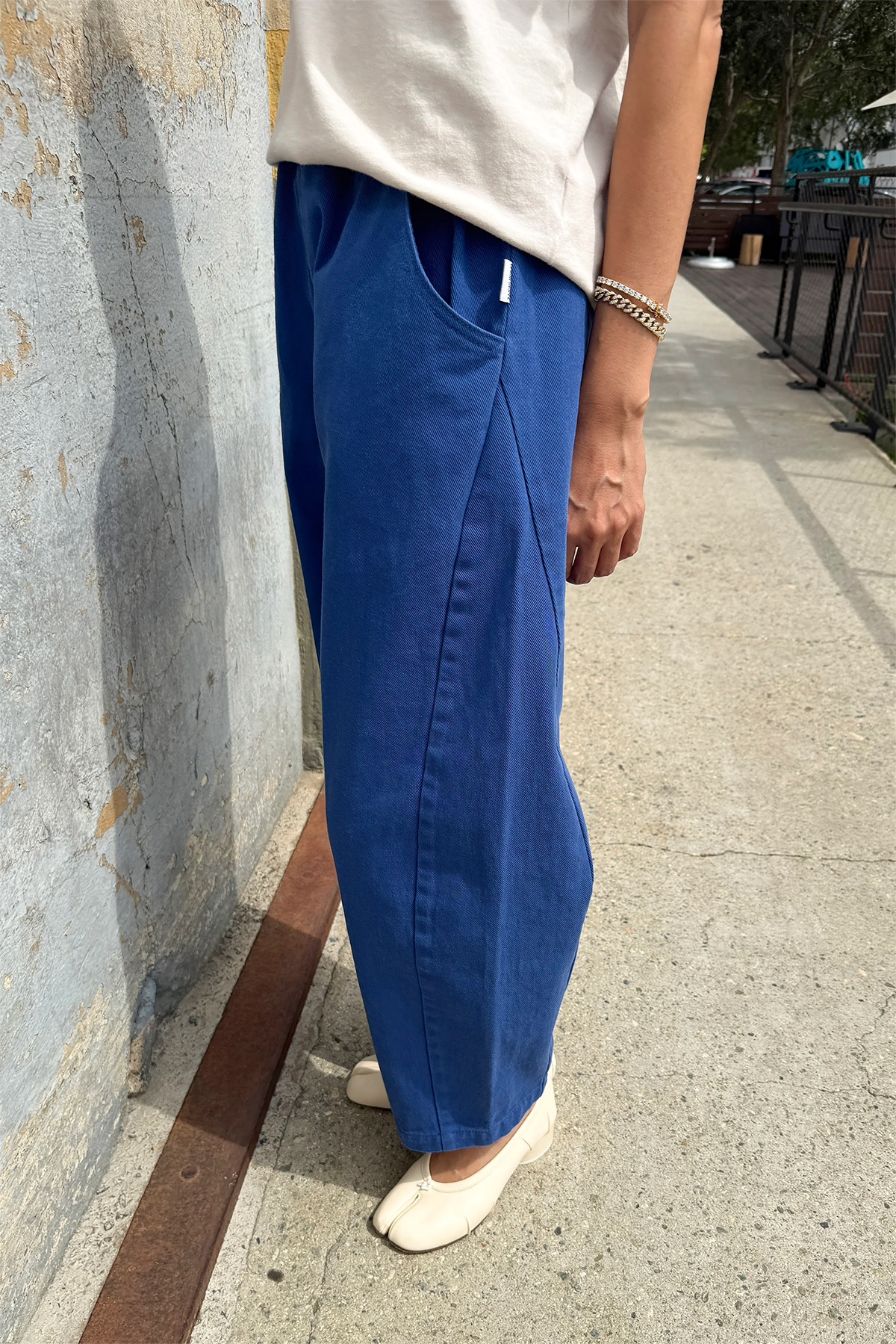 A person stands on a sidewalk wearing Le Bon Shoppe - Arc Pant / Azure, a high-waist, loose blue denim pant, paired with a white top and cream shoes; only the lower half is visible next to a textured, weathered wall.