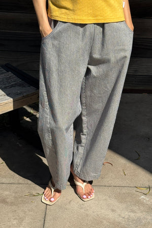 A person wearing Le Bon Shoppe - Arc Pant in Blue Stripe, paired with a yellow shirt and white sandals, stands outdoors on a sunny day with hands in pockets; only the lower body is visible.