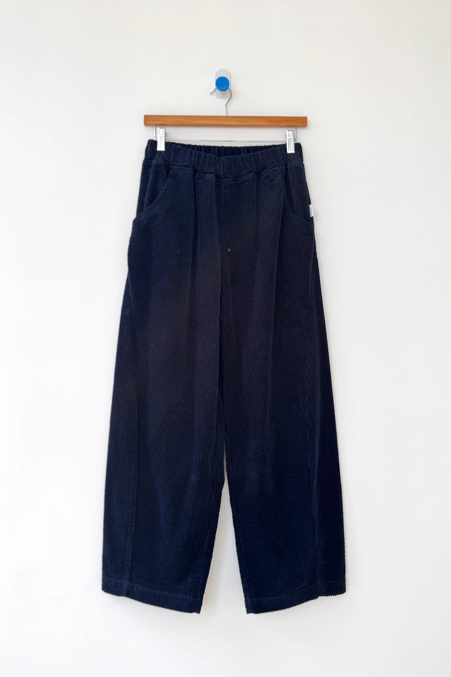Le Bon Shoppe - Arc Pant / Midnight Corduroy: Wide-leg, high-waist dark navy corduroy pants with an elastic waistband and side pockets, shown on a wooden hanger against a plain white wall.