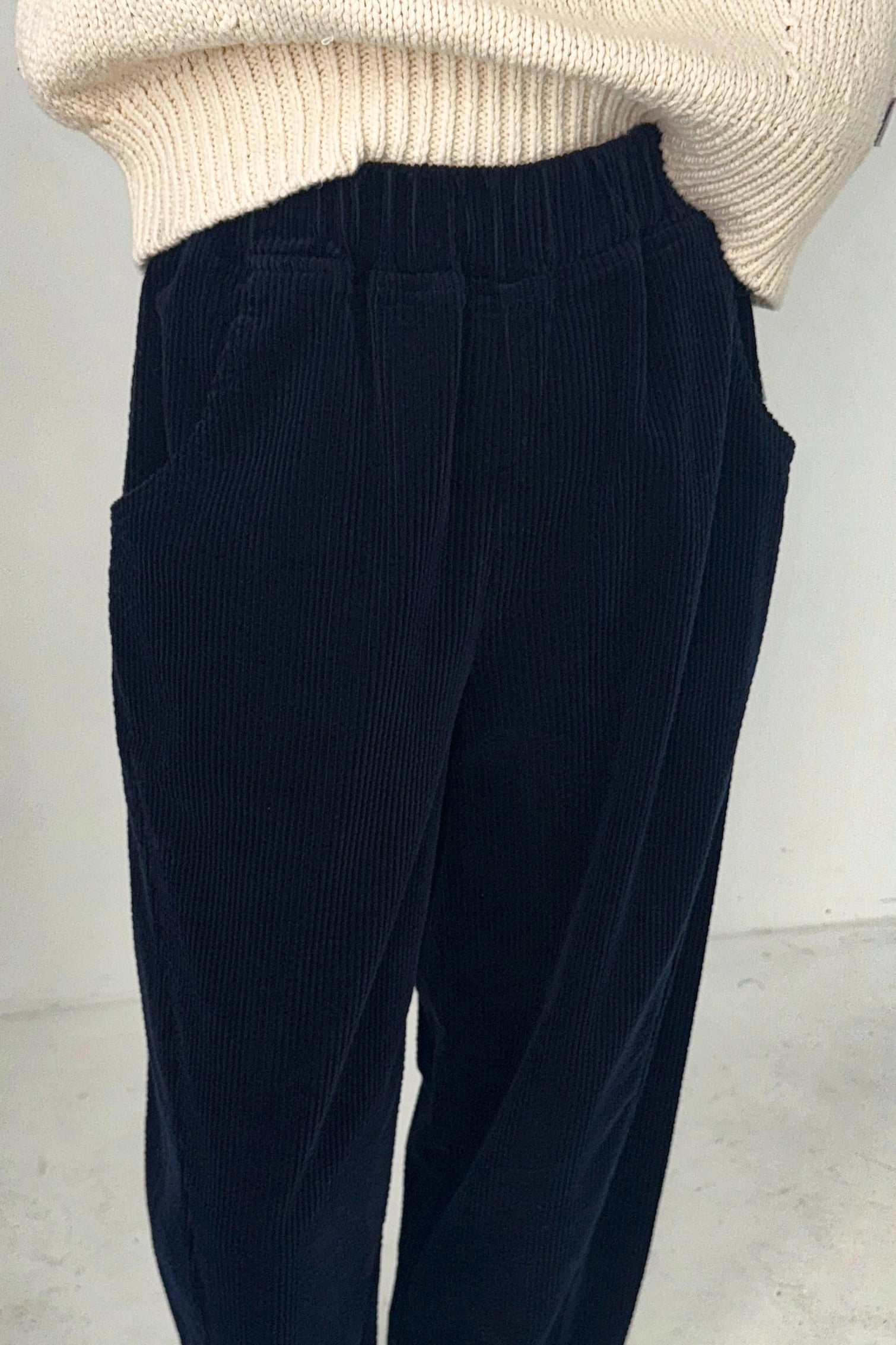 A person wears the Le Bon Shoppe Arc Pant in Midnight Corduroy—high-waisted, dark blue with side pockets and cropped legs—paired with a cream knit sweater against a light background.