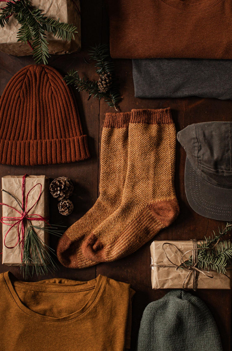 Flat lay of cozy winter clothing, including a rust beanie, mustard socks, green and brown shirts, a gray cap, and several wrapped gifts with pine branches and pinecones on a wooden surface.