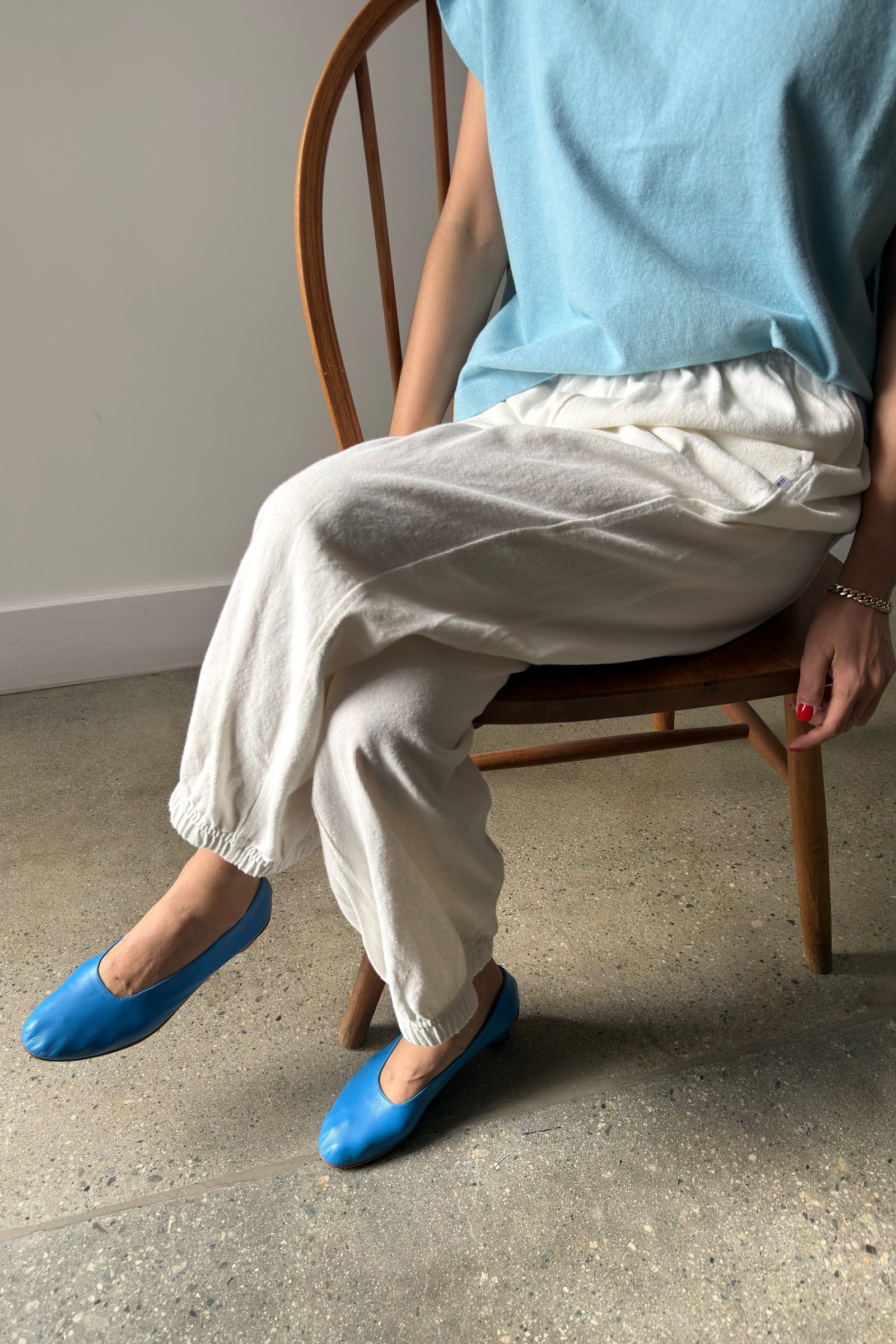 A person sits on a wooden chair wearing Le Bon Shoppe - Balloon Pant / White Cotton. Only their lower body and part of an arm are visible. The floor is concrete, and the background is plain.