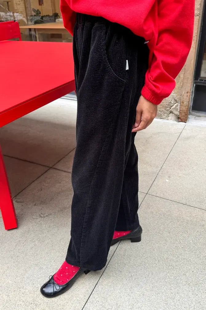 A person wears black Le Bon Shoppe - Soft Corduroy Arc Pant with a cropped, wide-leg fit and elastic high waist, paired with red socks and black ballet flats.