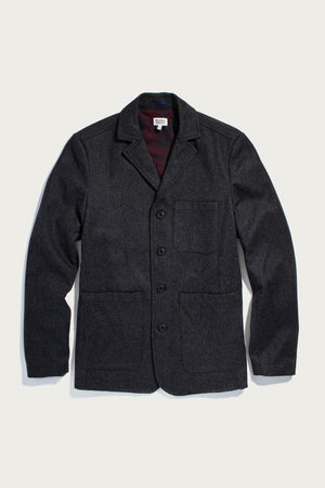 The Nate Chore Blazer in Onyx Wool by Bridge & Burn is a dark charcoal gray wool-blend blazer with a notched collar, button front, and three pockets. Laid flat on white, its dark red plaid lining adds a stylish winter touch.