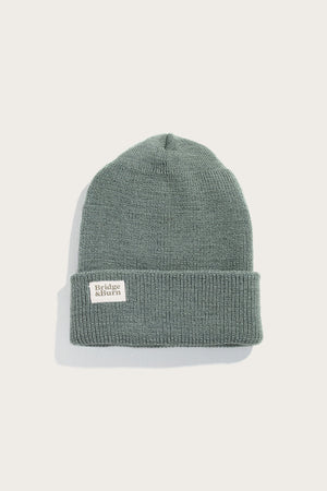 The Bridge & Burn Wool Watch Cap / Foliage is a sage green 100% wool beanie with a folded brim and a white rectangular brand patch on the front, shown against an off-white background.