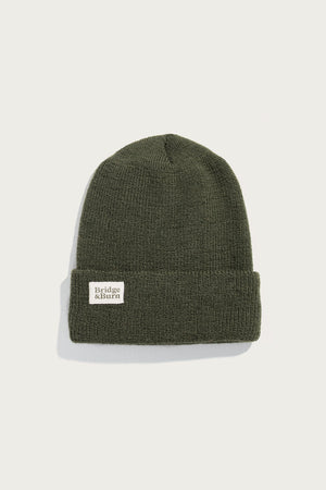 The Bridge & Burn Wool Watch Cap in Olive Drab is a 100% wool knit beanie with a folded brim and a white rectangular front patch. Made in the USA, it's shown against a plain white background.