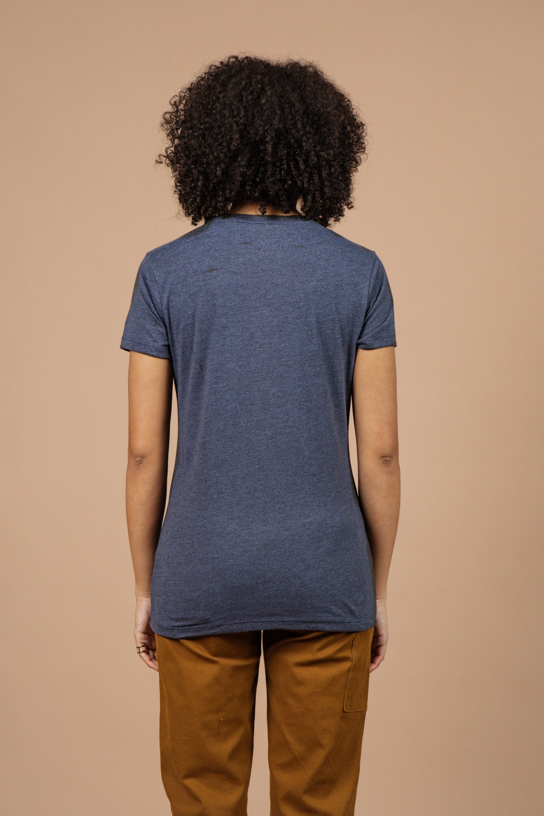 A person with curly hair, seen from behind, wears the Bridge & Burn Women's Alpenglow Tee in navy and brown pants against a plain beige background.
