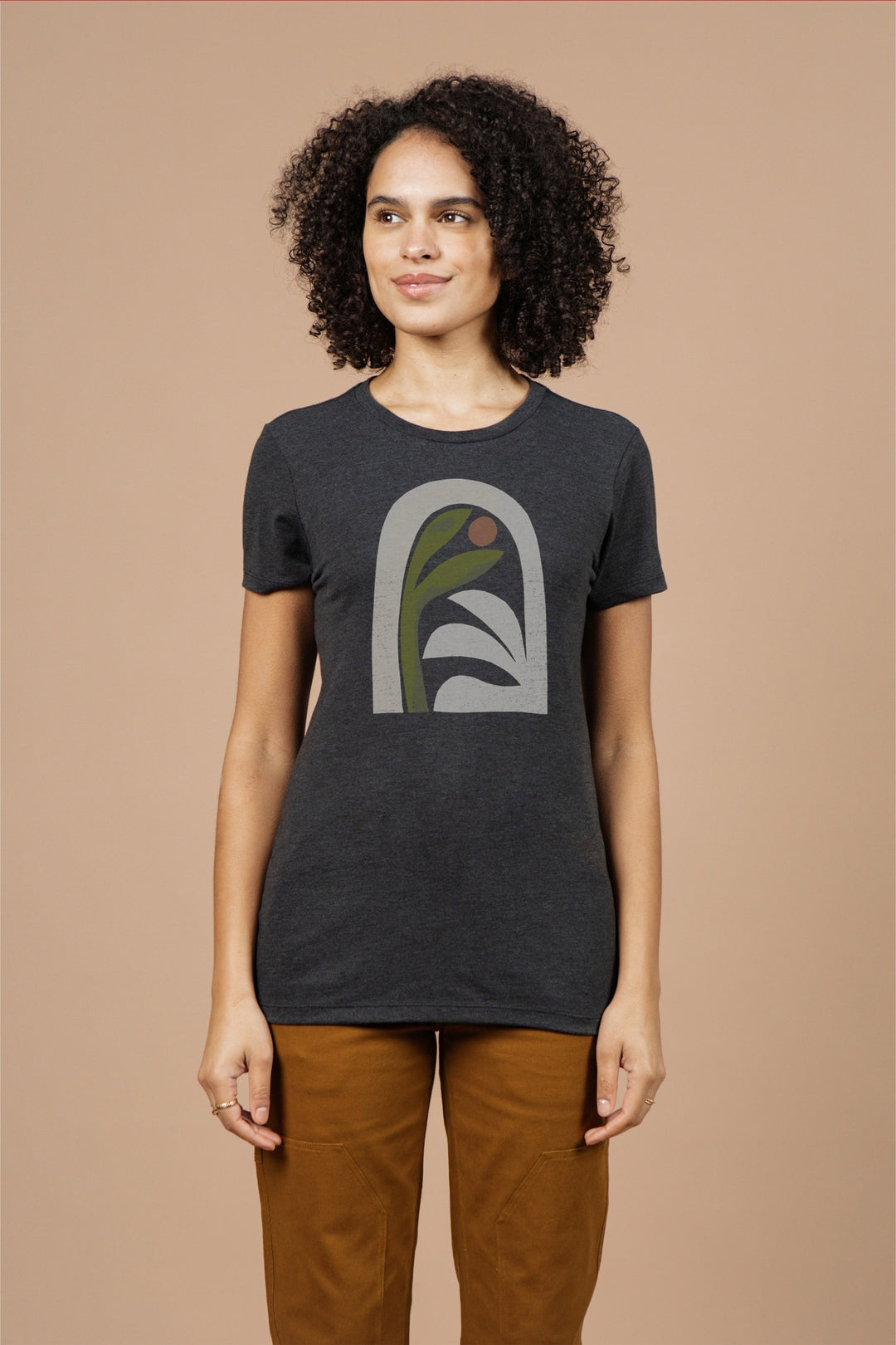 A woman with curly hair wears the Bridge & Burn Women's Arch Tee in Charcoal and brown pants, standing against a plain beige background.