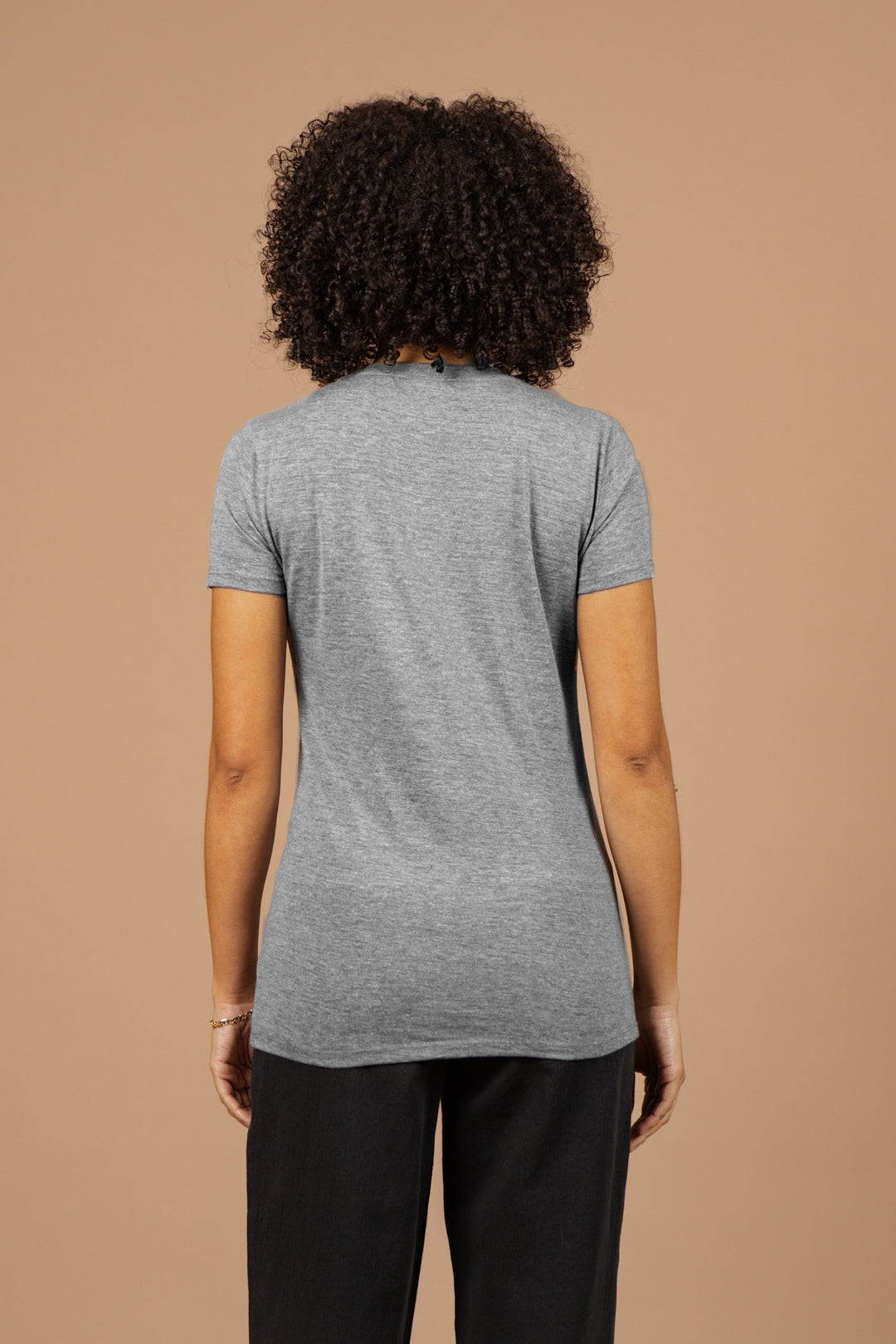 A person with curly hair faces away from the camera, wearing the Bridge & Burn Women's Cresting Wave Tee in grey and black pants against a plain beige background.