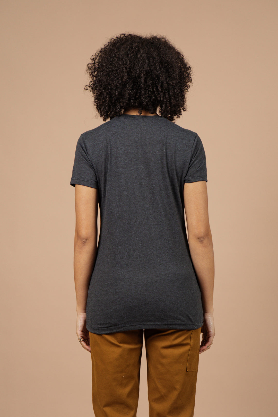 A person with curly hair faces away, wearing the Bridge & Burn Women's Night Watch Tee in black with brown pants, set against a plain beige background.