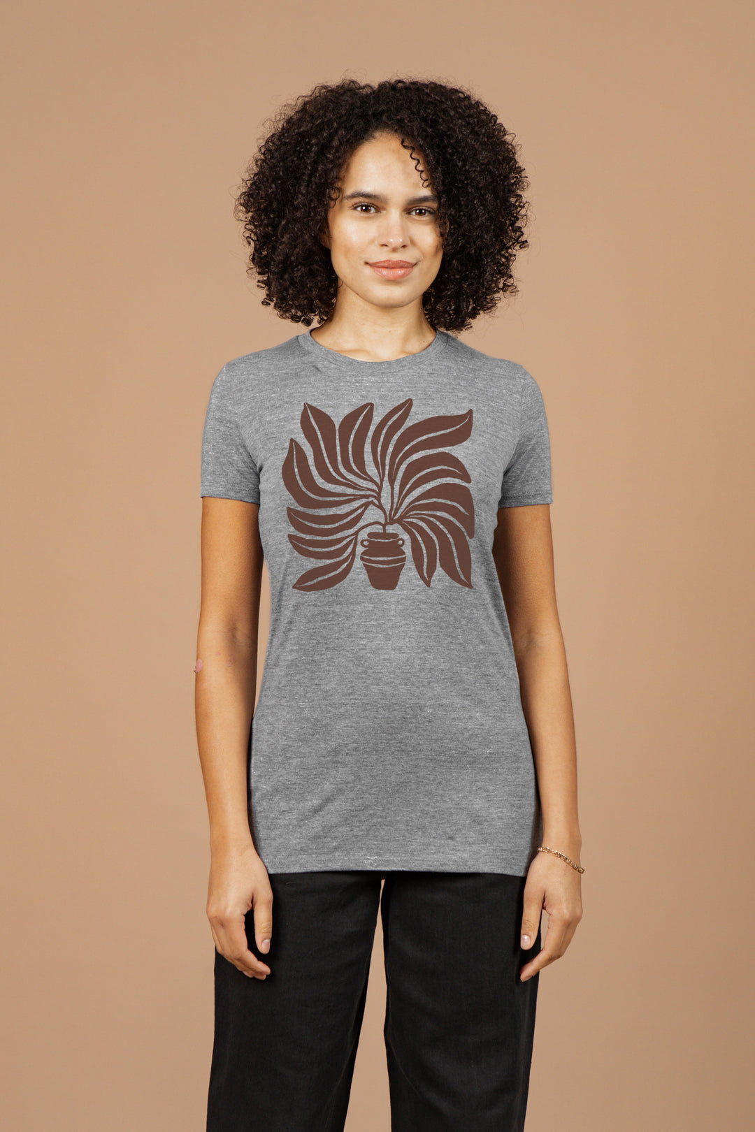 A woman with curly hair stands against a beige background, facing forward and smiling slightly in the Bridge & Burn Women's Grown Tee in grey, featuring organic cotton and a brown potted plant graphic, paired with black pants.