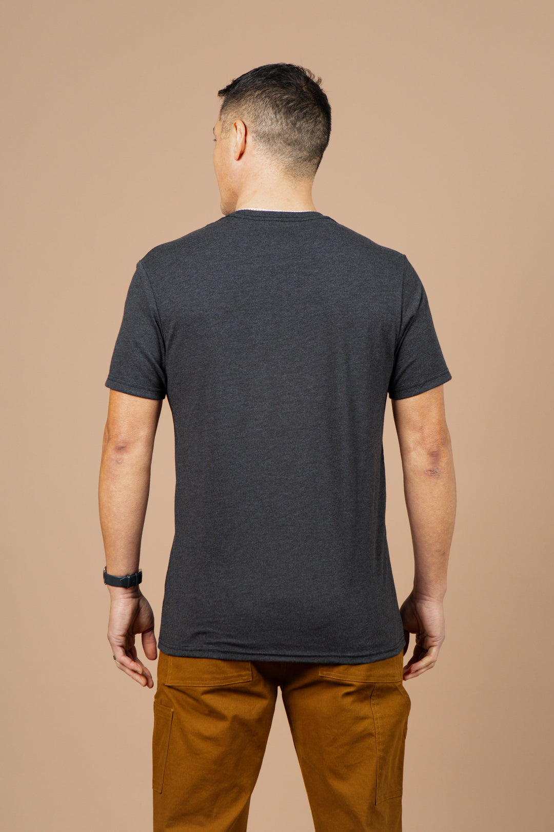 A man stands with his back to the camera, wearing the Bridge & Burn Men's Side Winder Tee in black and light brown pants against a plain beige background.