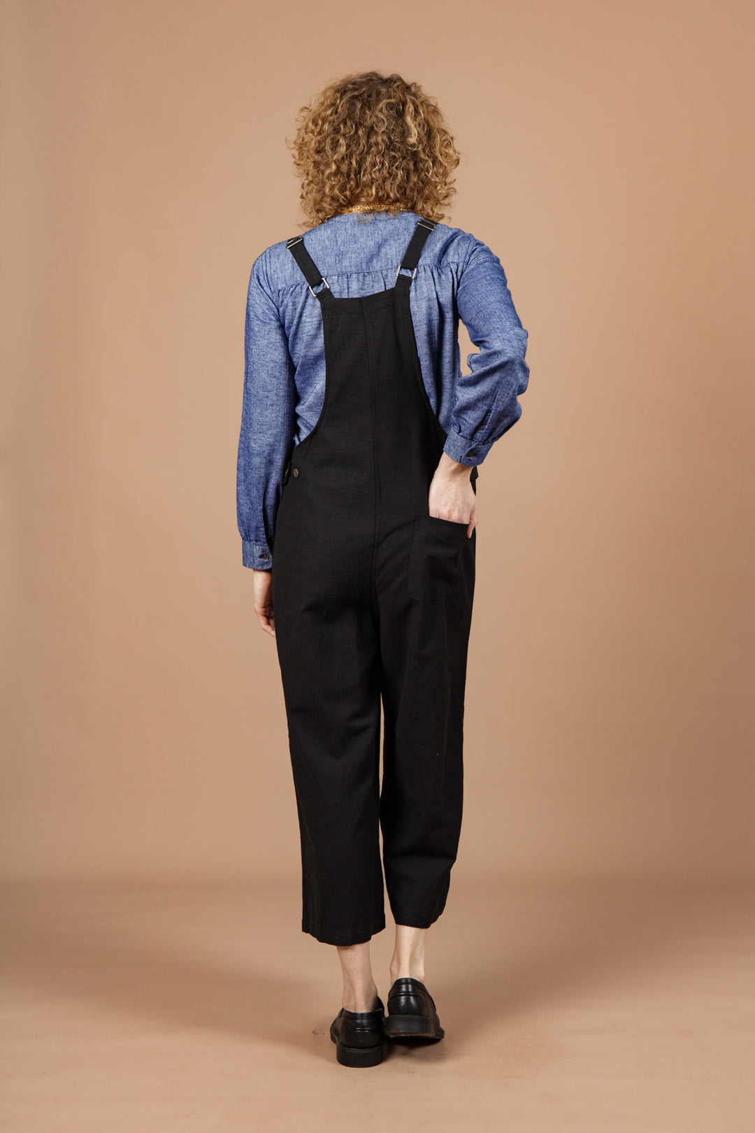 A person with curly hair, seen from behind, wears a blue long-sleeve shirt and Bridge & Burn's Edie Overall in black with adjustable straps, standing against a plain beige background.