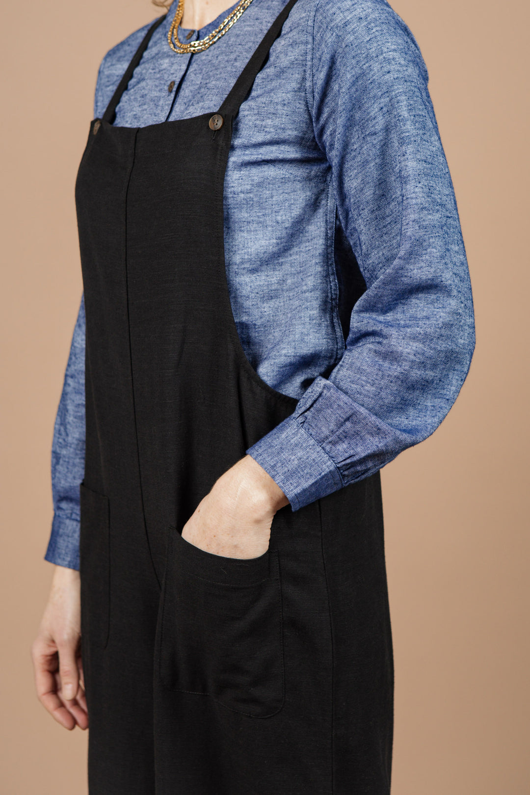 A person wears the Bridge & Burn Edie Overall in Black—a wide-leg, adjustable-strap style—over a blue long-sleeve shirt, standing with one hand in their pocket against a beige background. Their head is partially out of frame.