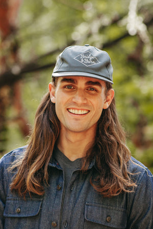 A smiling person with long brown hair stands outdoors against a blurred backdrop of greenery, wearing the Bridge & Burn Diamond Head Cap in grey—a cotton twill hat featuring a leather adjustment strap—and a blue button-up shirt.
