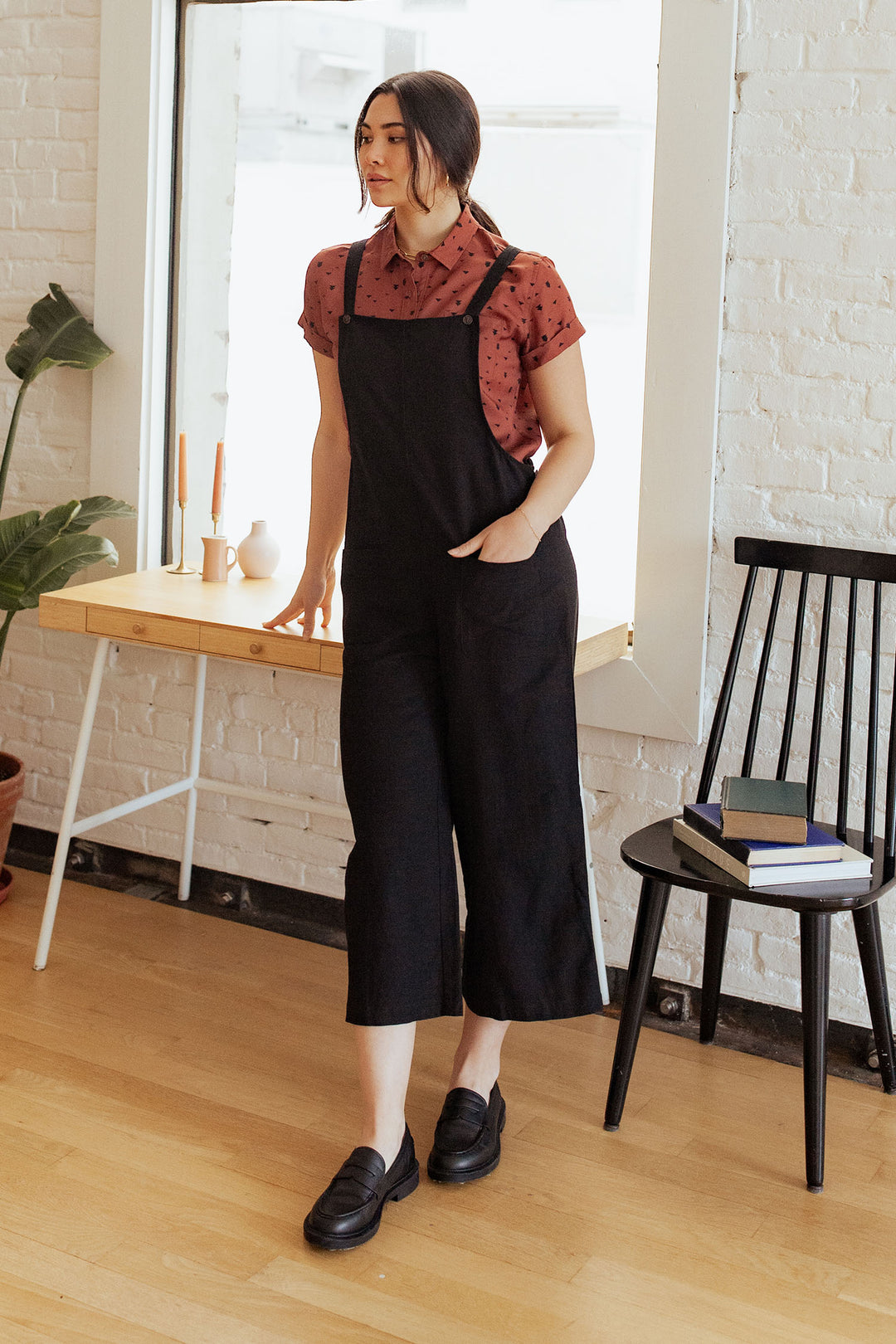 A woman stands indoors in Bridge & Burn's Edie Overall / Black—wide-leg tencel-cotton overalls with adjustable straps—paired with a maroon tee and black shoes, near a wooden desk, vases, a plant, and a book-filled black chair.