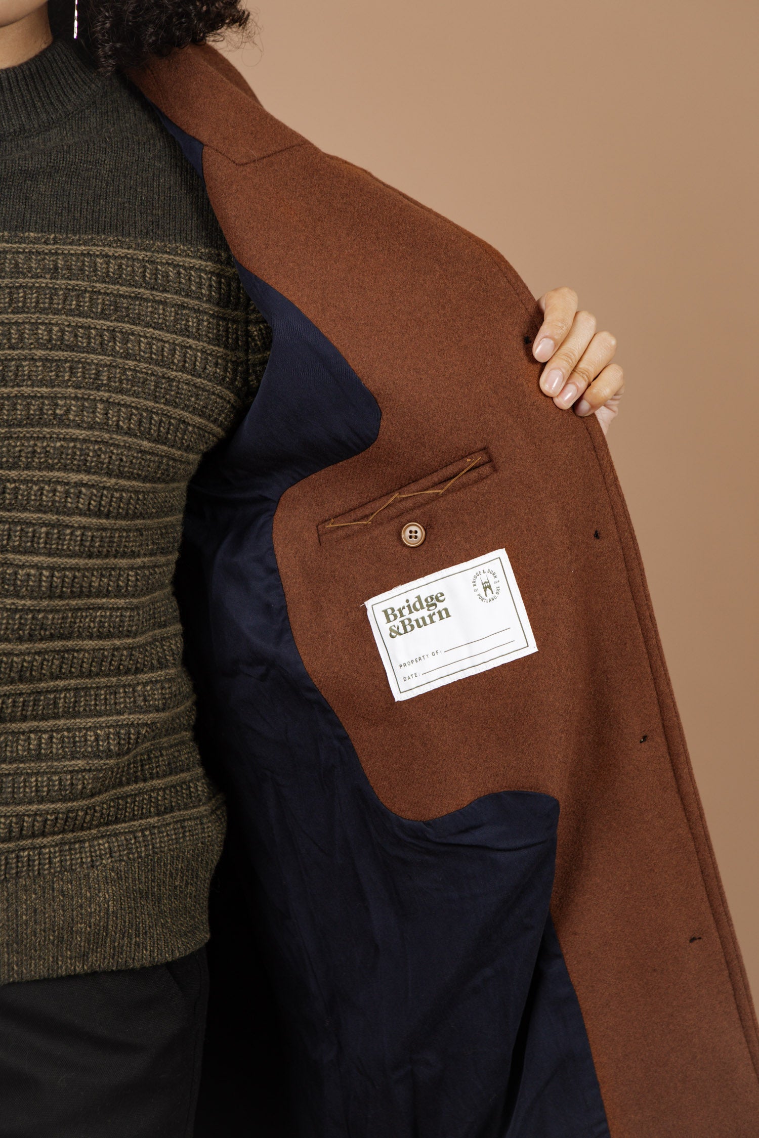 A person in a green knit sweater opens the Alberta Car Coat in Fired Brick by Bridge & Burn, revealing its dark lining and inner pocket with a white brand label. The background is beige.