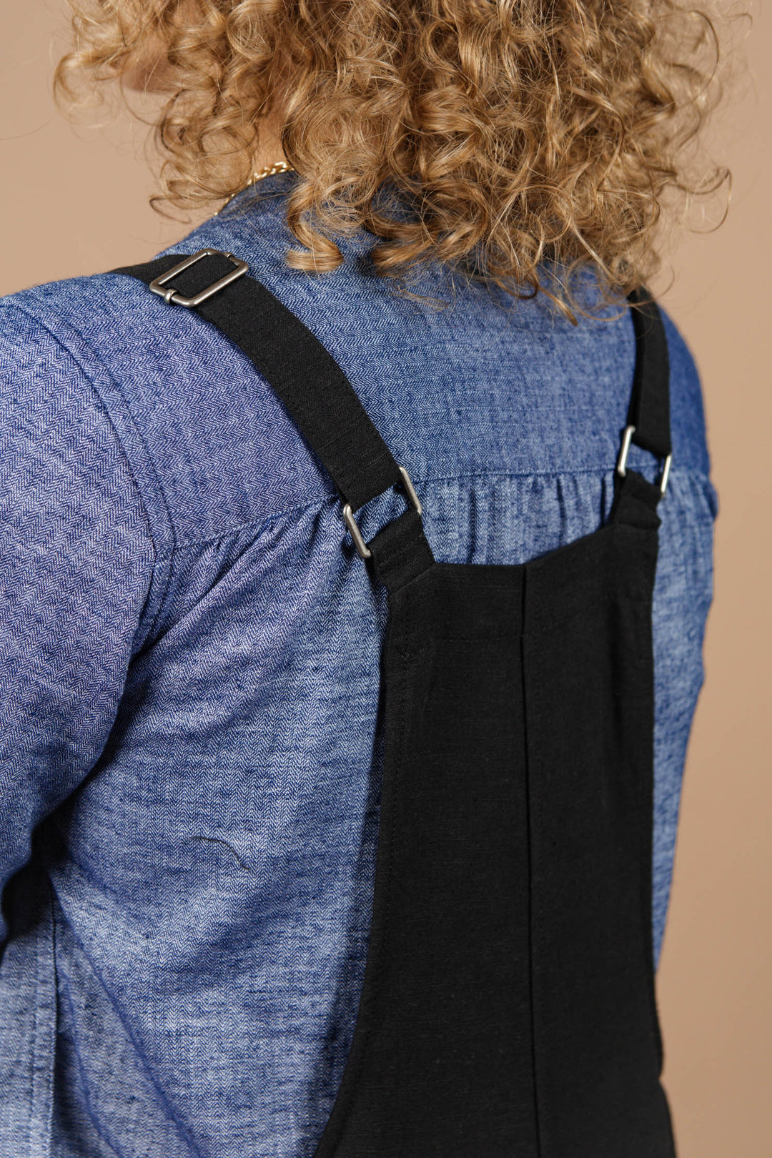 Close-up of a person with curly hair wearing the Bridge & Burn Edie Overall in Black, styled with a blue denim shirt. The overalls feature adjustable straps at the back and reveal part of the upper back.