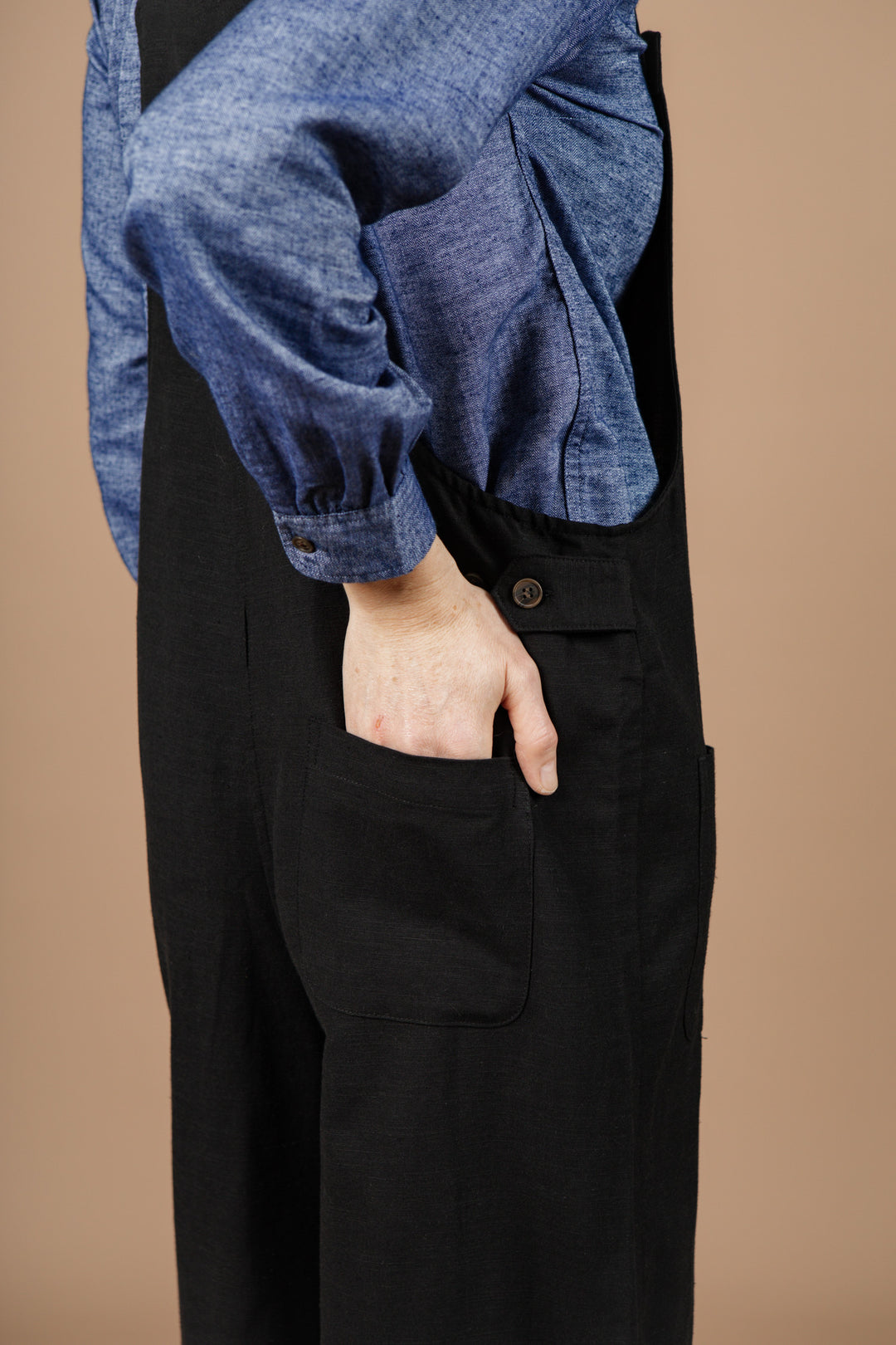 A person wearing the Bridge & Burn Edie Overall in black, paired with a blue long-sleeve shirt, stands against a neutral brown background with their right hand tucked into the pocket.