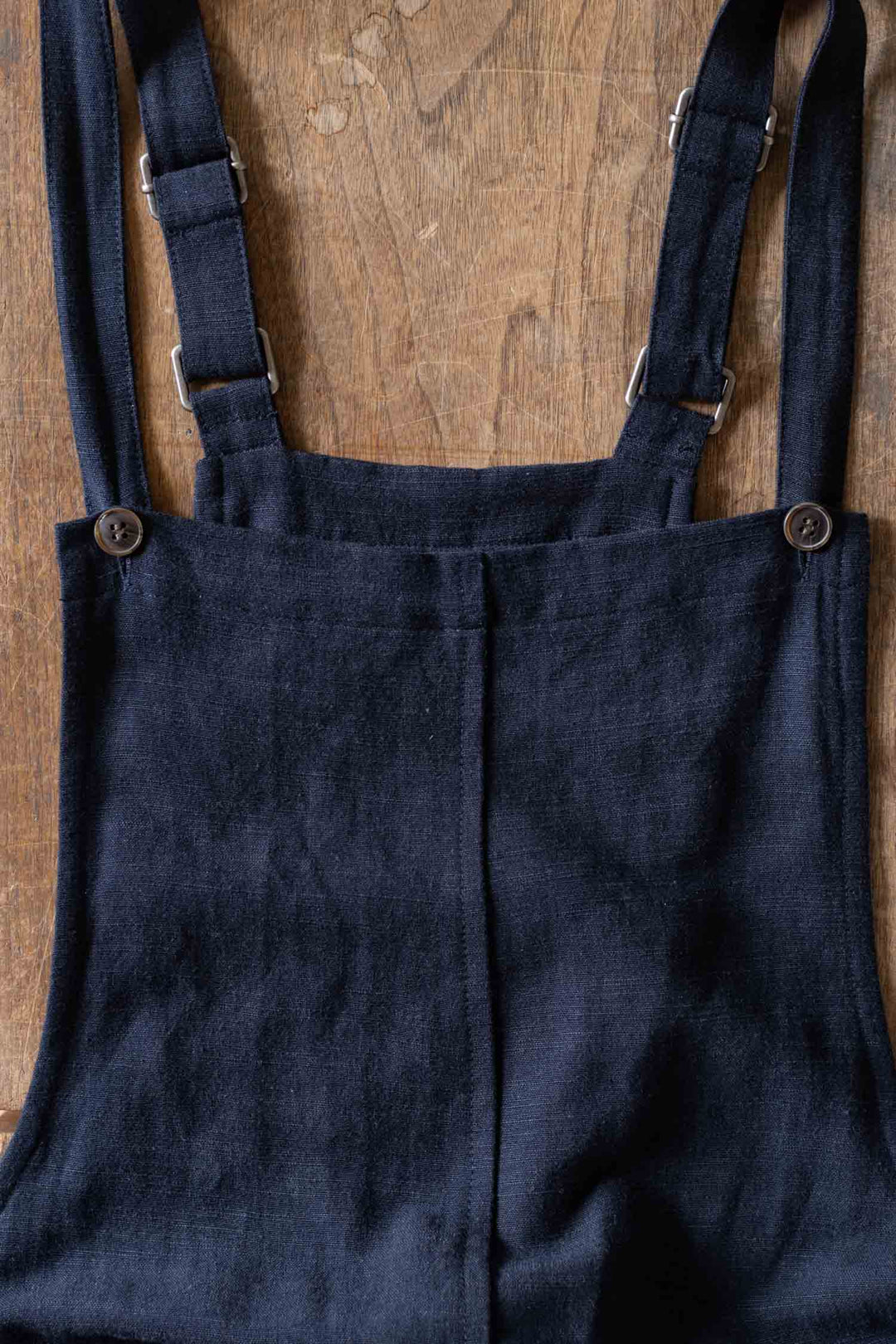 The Edie Overall in Navy by Bridge & Burn, made from a soft linen-cotton blend with adjustable straps and top buttons, is shown laid flat on wood with only the upper portion visible.