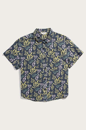The Bridge & Burn Greer Cropped Shirt in Spring Kelp features a dark base with yellow-green and light purple botanical prints, a boxy fit, collar, short sleeves, and a breathable cotton-linen blend for comfort.