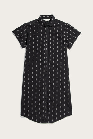 The Loren Shirt Dress in Onyx Arrows by Bridge & Burn is a short-sleeved, knee-length black cotton dress with a collar, subtle white vertical pattern, button front, and curved hem—ideal for versatile workwear.