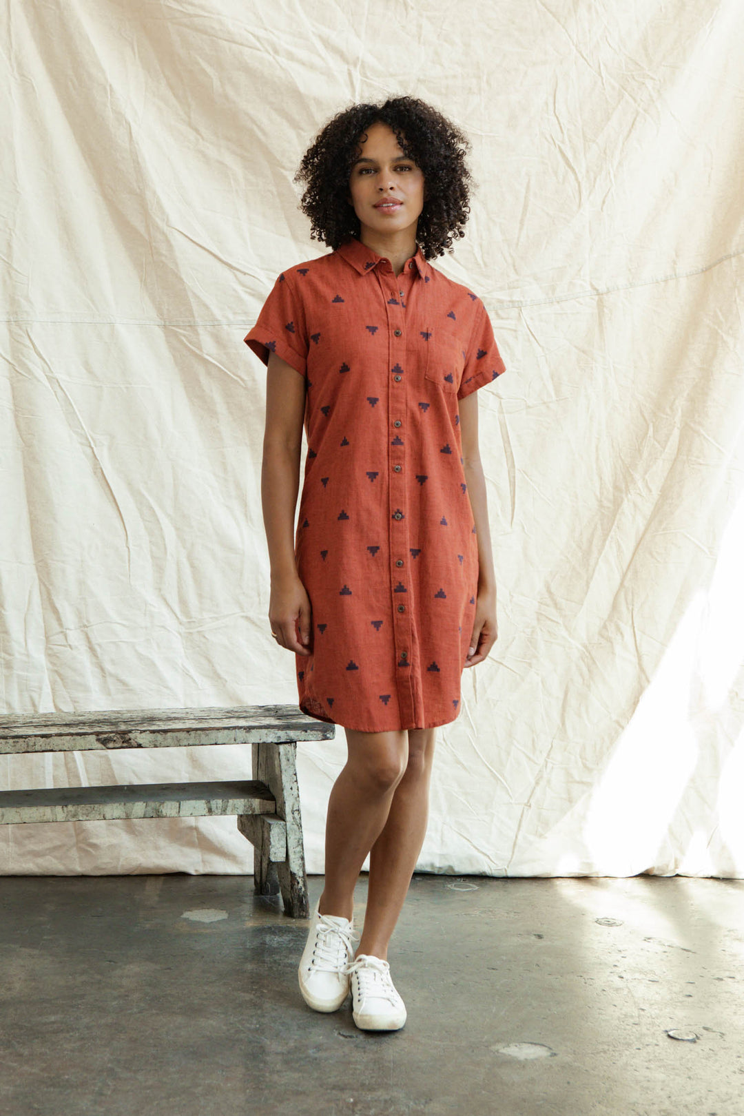 A woman stands on a concrete floor indoors, posing in front of a light beige fabric backdrop and wooden bench. She wears the Bridge & Burn Loren Shirt Dress in Rust Fil-Coupé Pyramids with white sneakers.