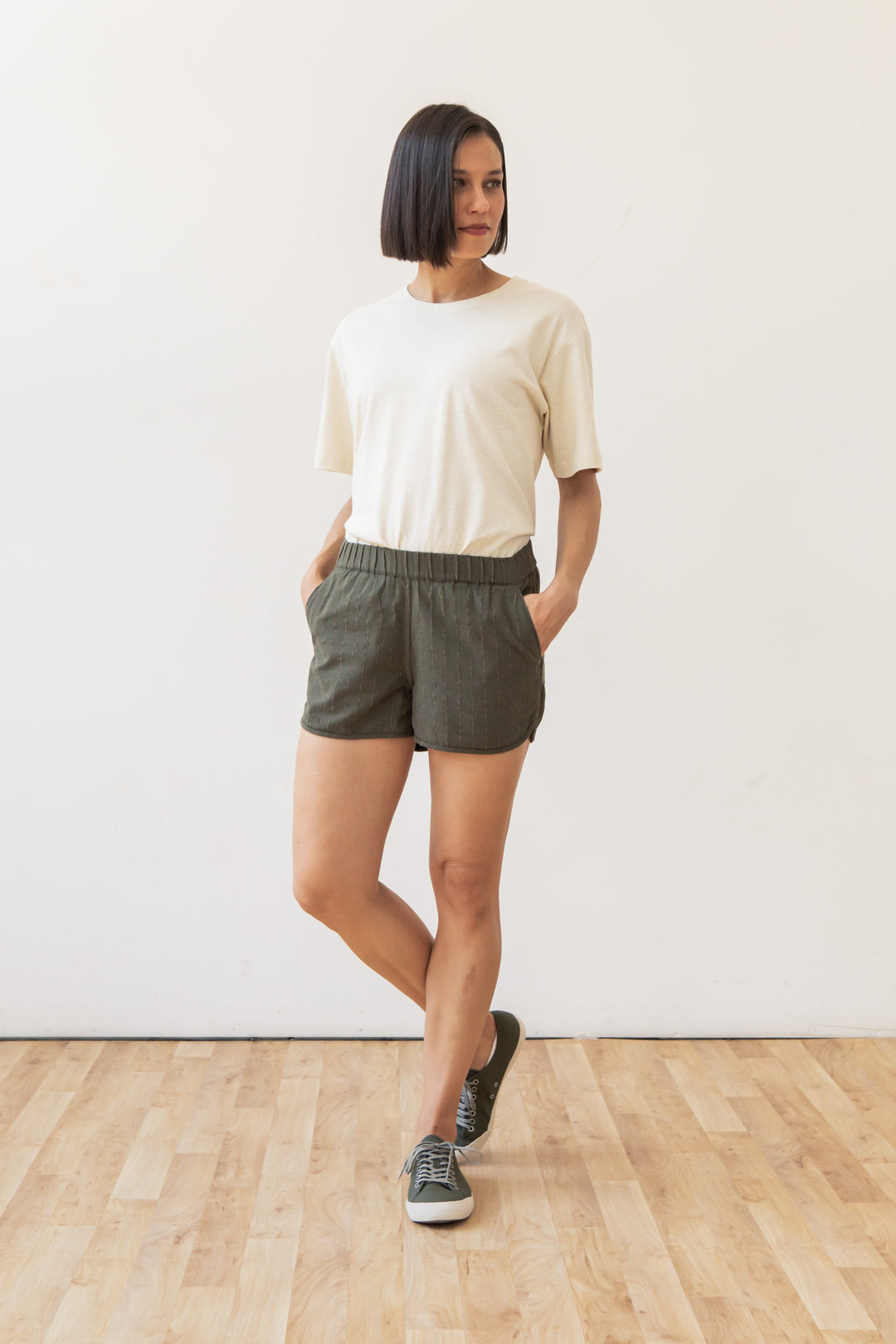A woman with a short bob stands on a wooden floor, wearing a white tee, Bridge & Burn's Luca Track Short in Olive Stripe, and gray sneakers. Hands in pockets, she poses casually and looks slightly to the side.